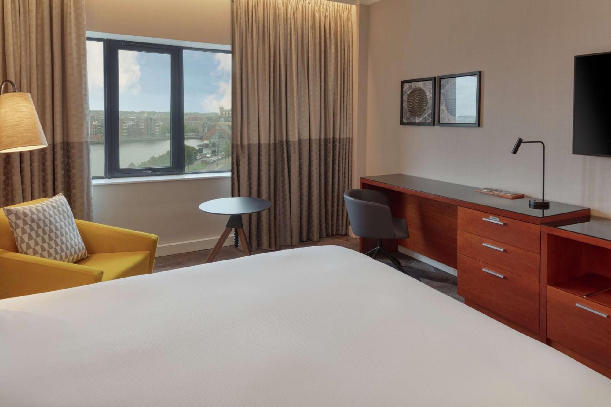 فندق هيلتون بيلفاست بلفاست المظهر الخارجي الصورة The photo shows a modern hotel room. It features a large bed with a plain white bedspread, and there's a stylish yellow armchair next to it. A small round table is placed nearby, along with a gray chair. The room has large windows covered with light 