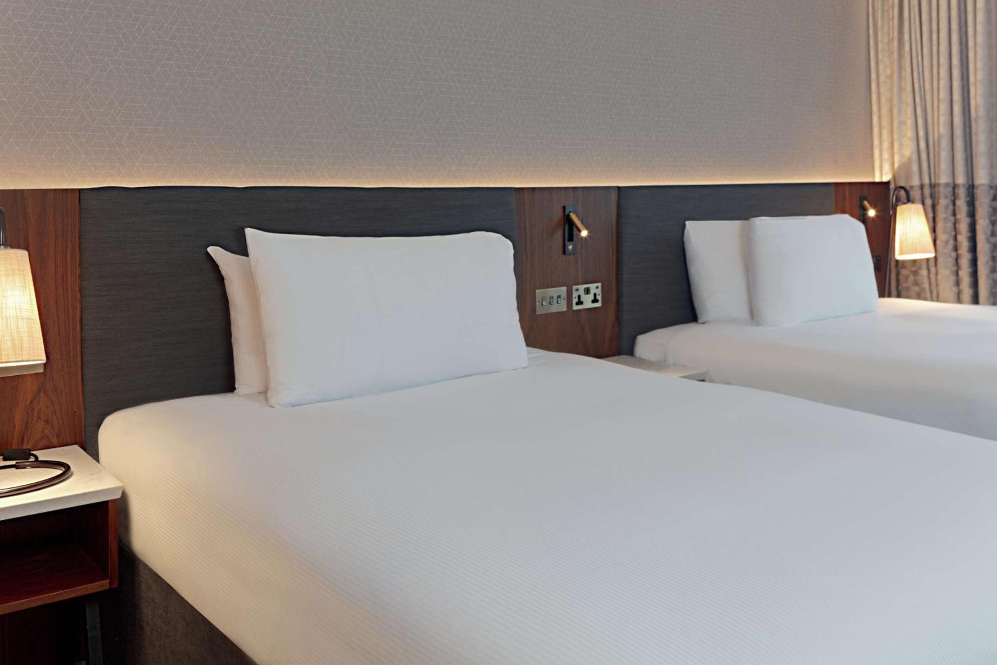 فندق هيلتون بيلفاست بلفاست المظهر الخارجي الصورة The photo shows a modern hotel room with two neatly made beds. Each bed features white linens and pillows, with a simple yet stylish headboard in a dark wood finish. The room has a soft, ambient light illuminating the area, creating a welcoming atmos