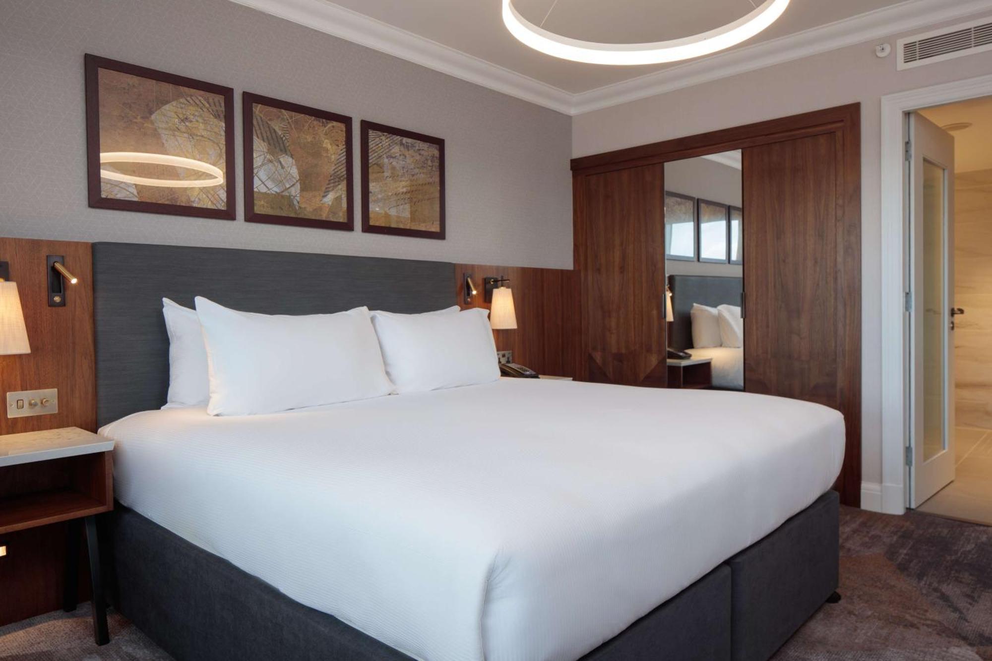 فندق هيلتون بيلفاست بلفاست المظهر الخارجي الصورة The photo shows a modern hotel room with a clean and contemporary design. The focal point is a large bed with crisp white bedding and two fluffy pillows. The headboard is gray, and next to the bed, there are stylish bedside lamps. Above the bed, ther