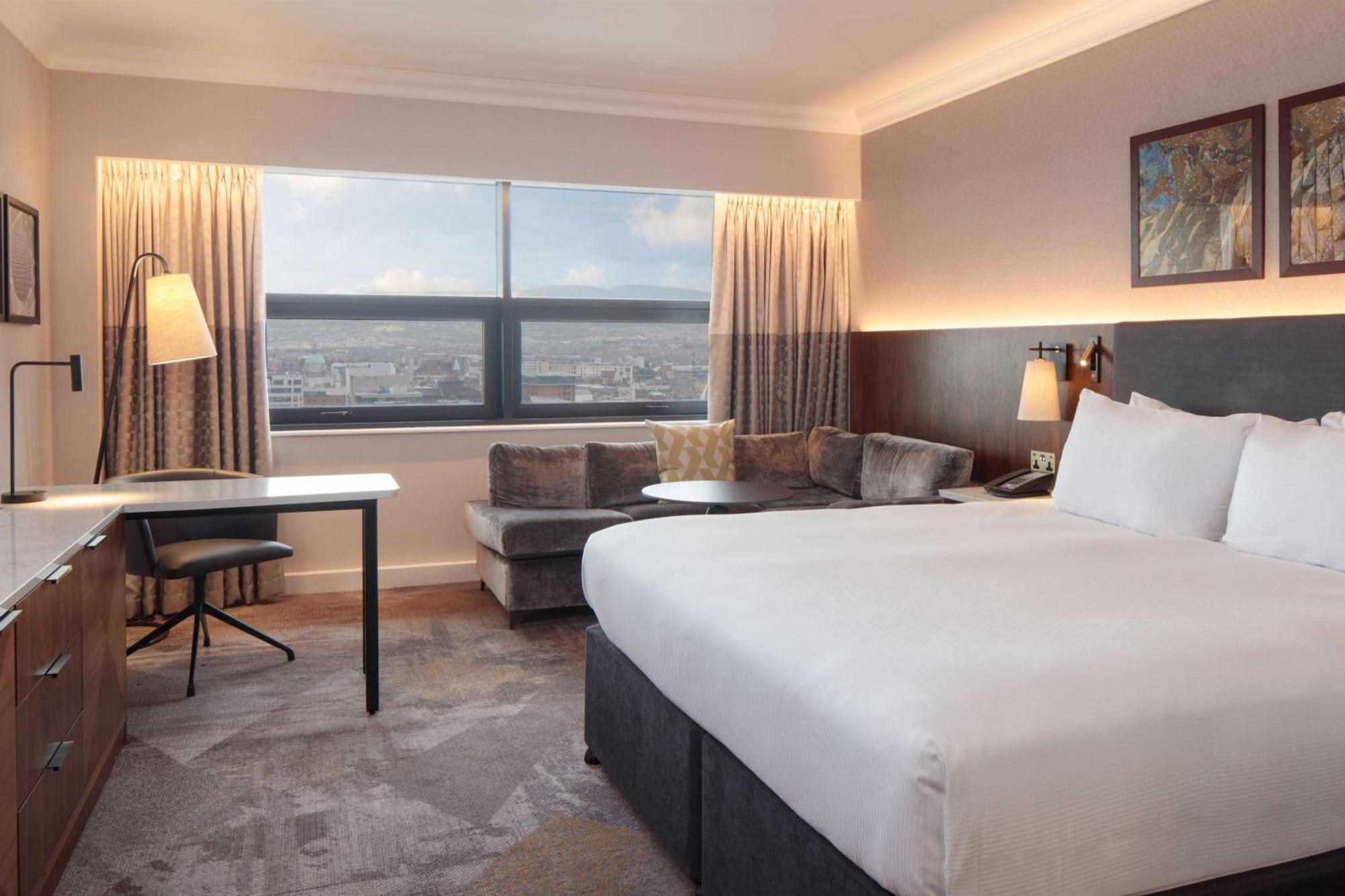 فندق هيلتون بيلفاست بلفاست المظهر الخارجي الصورة The photo shows a modern hotel room featuring a large bed with white linens positioned against a wall. On one side, there is a small seating area with a couch and a round table. A desk and chair are also present, suggesting a workspace. The room has 