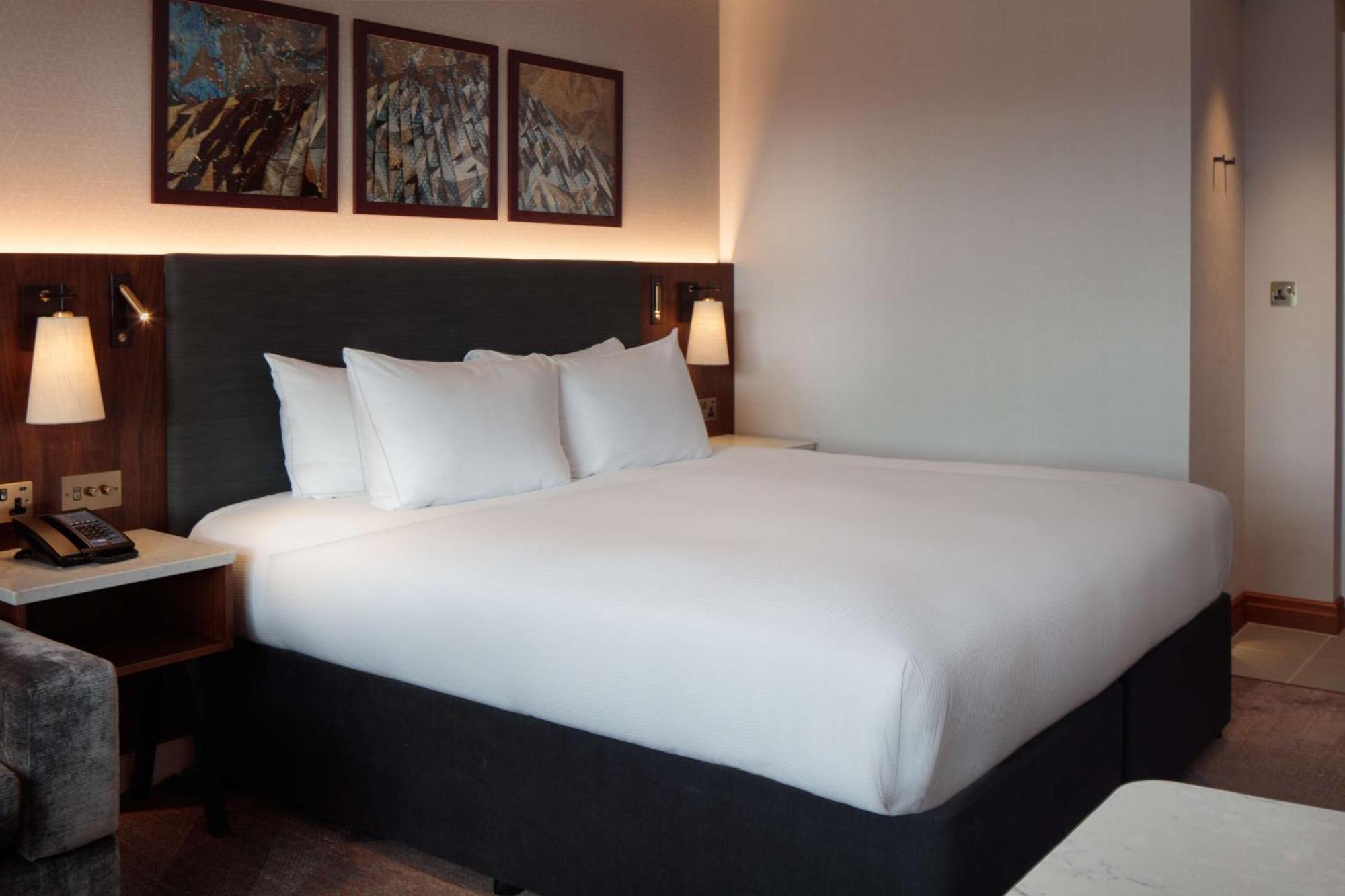 فندق هيلتون بيلفاست بلفاست المظهر الخارجي الصورة The photo shows a modern hotel room featuring a large bed with white bedding and several pillows. The bed is positioned against a dark-colored headboard. On the wall above the bed, there are three framed artworks. The room has soft lighting, and ther