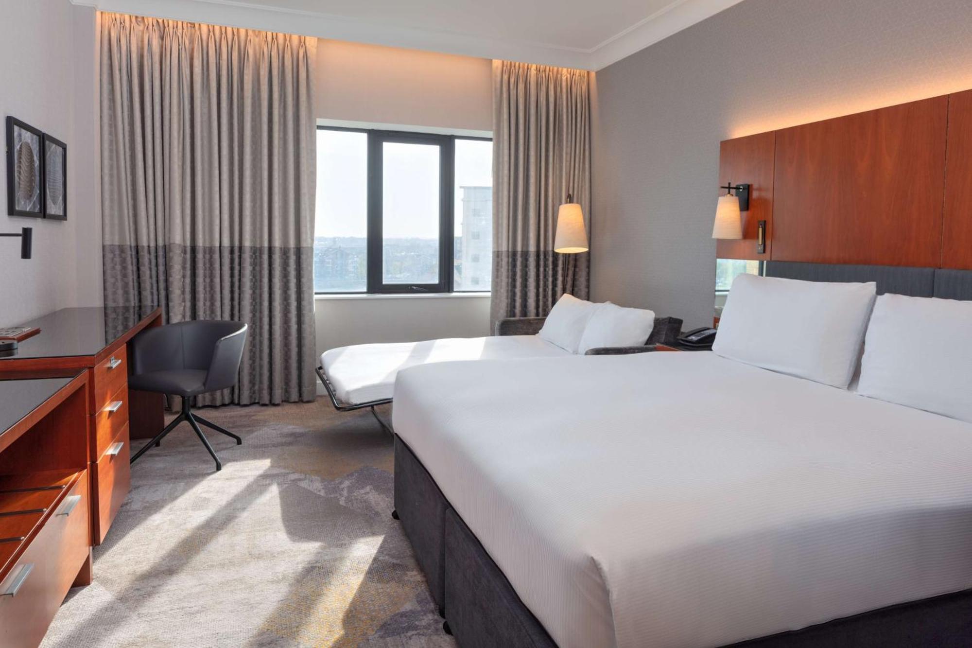 فندق هيلتون بيلفاست بلفاست المظهر الخارجي الصورة The photo shows a modern hotel room. It features two beds with white linens and a simple, elegant design. There is a large window allowing natural light to enter, covered by light-colored curtains. A comfortable chair and a small desk are placed near