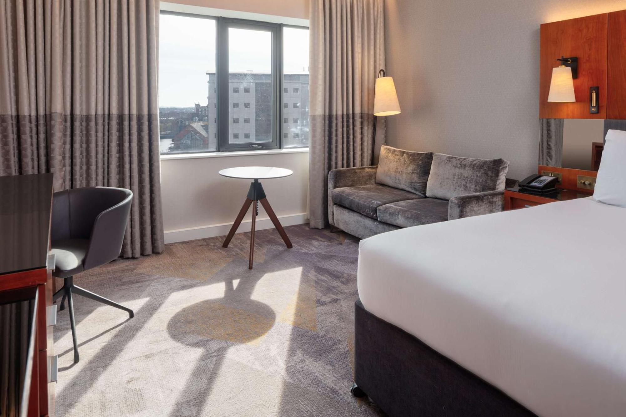 فندق هيلتون بيلفاست بلفاست المظهر الخارجي الصورة The photo shows a hotel room featuring modern furnishings. There is a large bed with white bedding positioned against one wall. Near the window, which has light-colored curtains, there is a small round table with a tripod-style base. A sleek gray sof