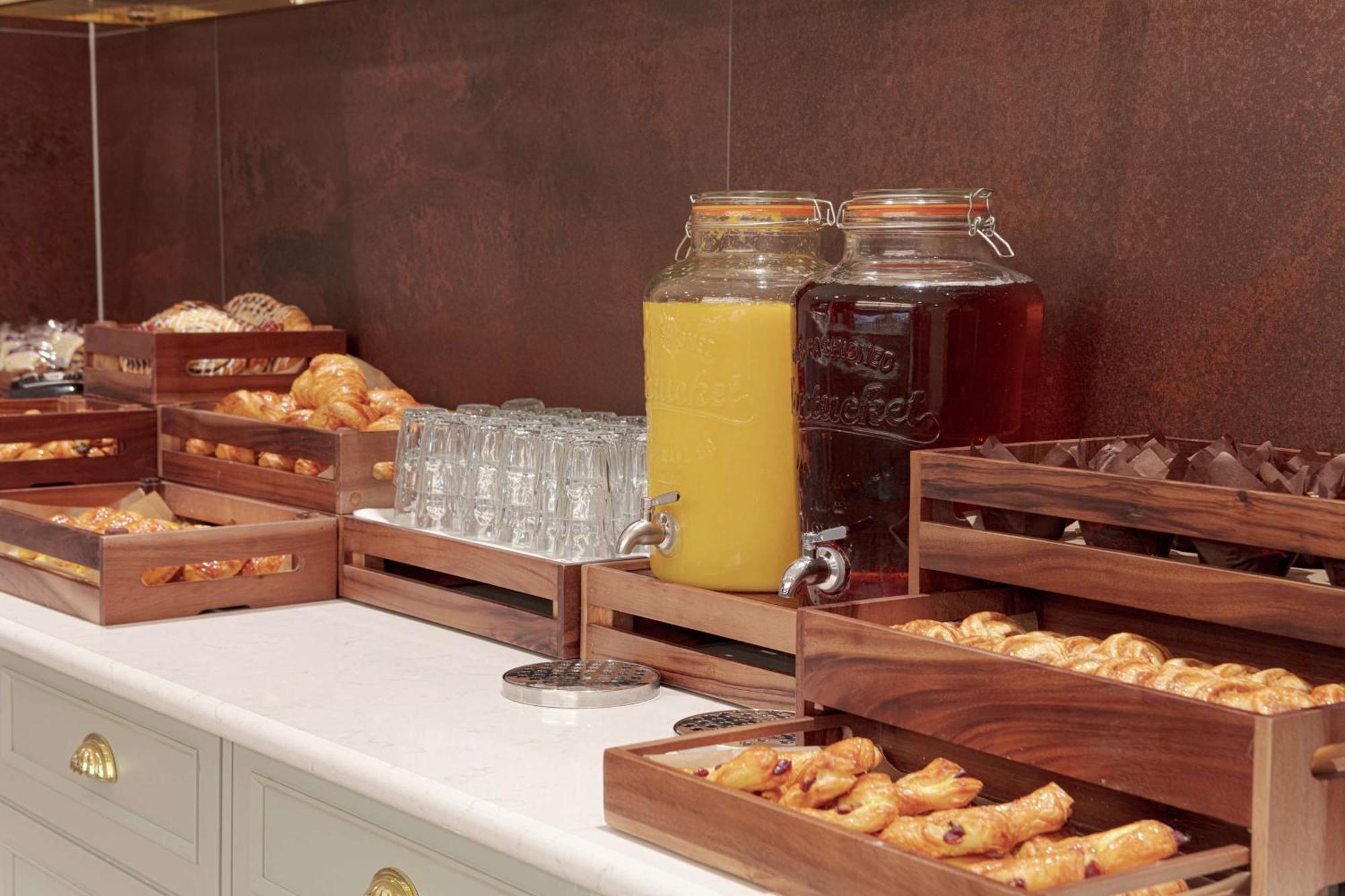 فندق هيلتون بيلفاست بلفاست المظهر الخارجي الصورة The photo shows a buffet-style setup featuring various food and beverage items. There are several wooden trays filled with pastries, including croissants and other baked goods. In the background, there are two large glass jars, one filled with yellow