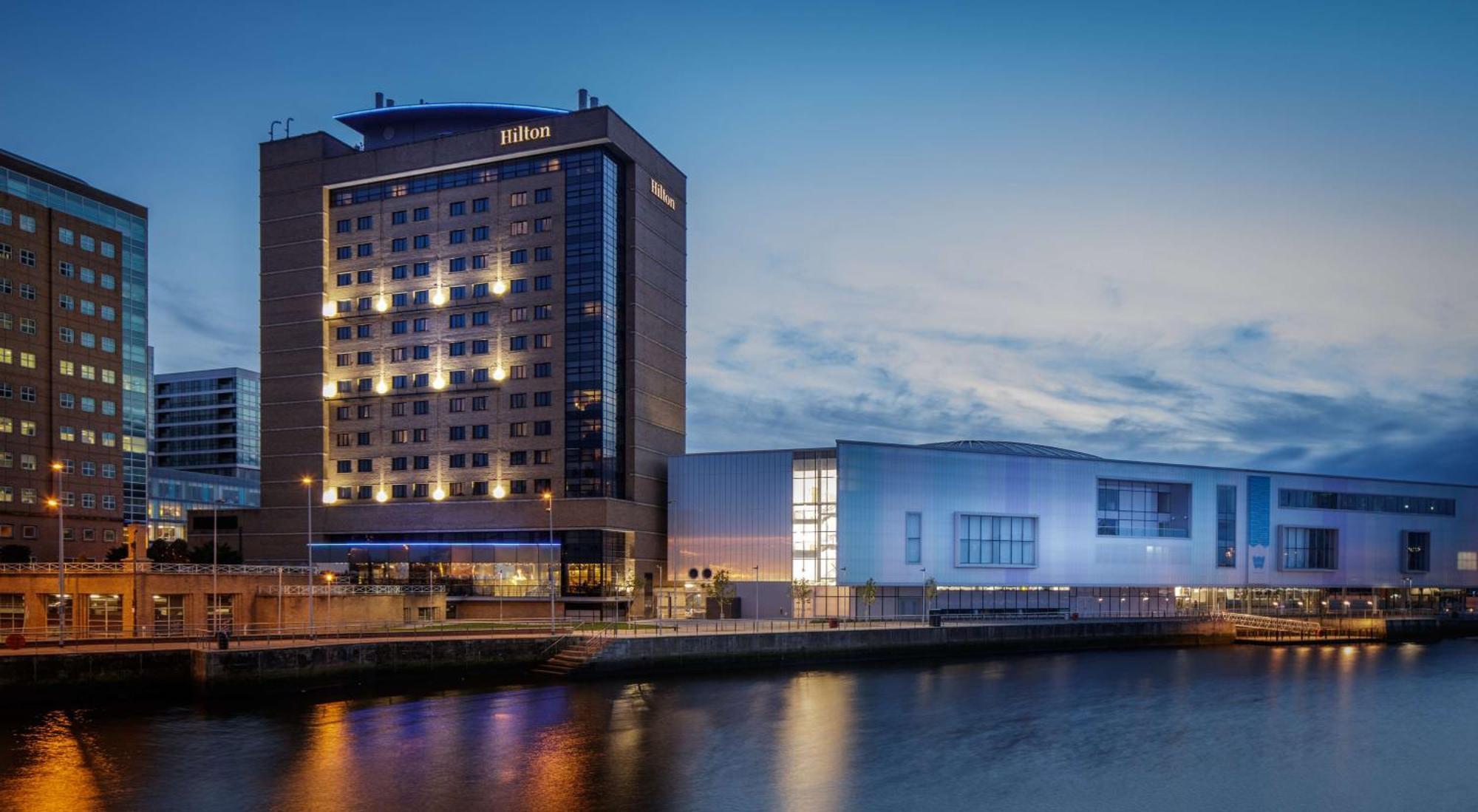 فندق هيلتون بيلفاست بلفاست المظهر الخارجي الصورة The image shows a Hilton hotel building along a waterway, illuminated during twilight. The hotel features a modern architectural design with multiple stories and large windows. To the right of the hotel, there is another structure that appears to be 